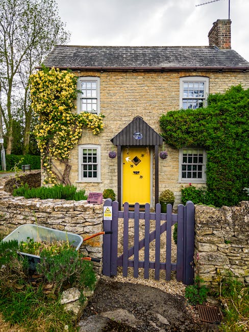 A charming two-story stone cottage featuring a vibrant yellow front door with black diamond-shaped decorations, situated behind a small purple picket gate. The front garden showcases a variety of well