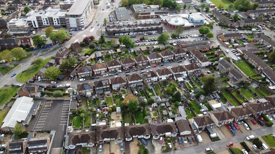 An aerial view of a suburban residential area showing a row of terraced houses with small, well-maintained gardens featuring lush green lawns, trimmed hedges, and mature trees. Some gardens have flowe